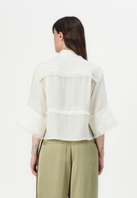 Blouse blanche raccourcie avec des détails en franges, des manches larges et une texture légère. Associe avec un pantalon vert olive, vue de derrière.