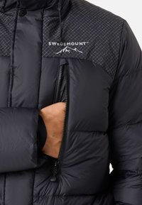 Black padded jacket with a geometric patterned upper, featuring a zippered pocket on the chest and a logo. Soft texture, warm design.