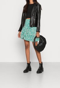 Frau trägt ein schwarzes Crop-Top, eine schwarze Lederjacke, einen grünen Blumenkleid und schwarze Stiefeletten, während sie eine schwarze gesteppte Handtasche hält.