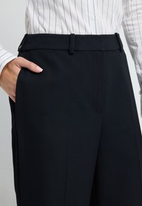 Czarne spodnie o teksturowanej tkaninie, z płaskim przodem, kieszeniami po bokach i gładkim paskiem. Połączone z białą koszulą w paski.