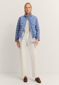 Hellblaues Steppjacke mit einer gesteppten Textur, kombiniert mit einem weißen gestreiften Hemd und weißen weiten Hosen. Braune Loafers runden das Outfit ab.