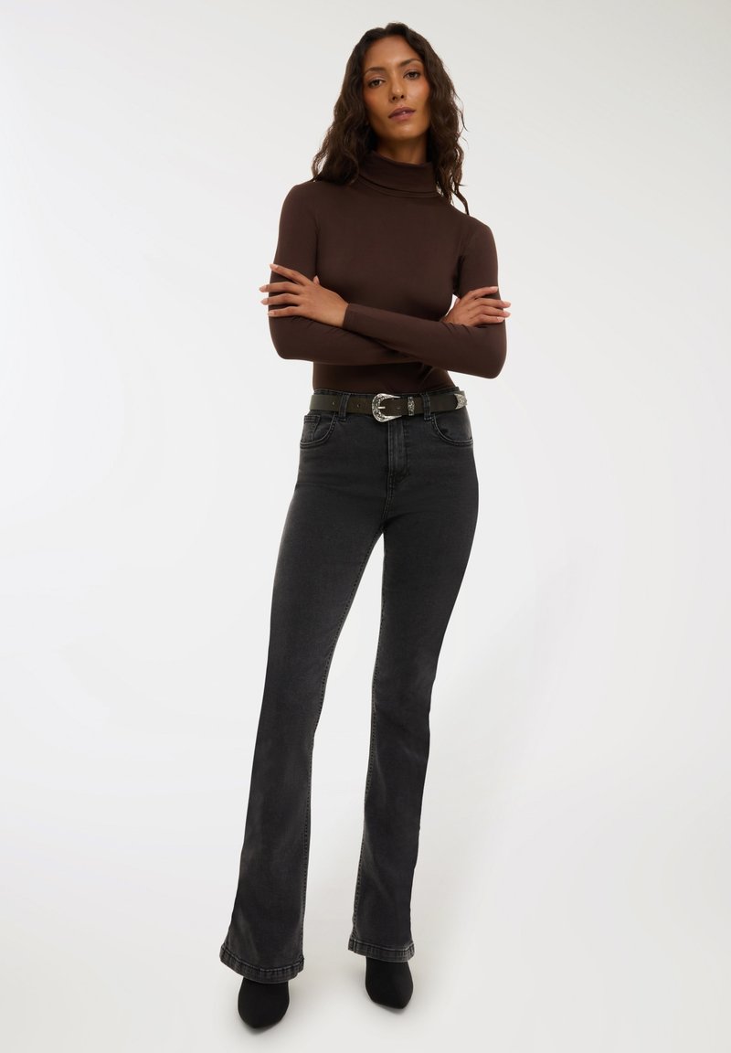 Kobieta z długimi kręconymi włosami, ubrana w brązowy golf, czarne rozszerzane dżinsy, czarny pasek i czarne szpiczaste buty, stoi z założonymi rękami.