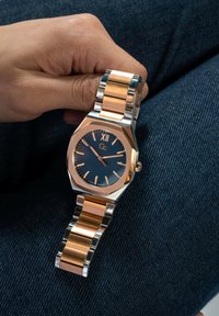 Rankose, laikrodžio su dviem spalvomis metaliniu korpusu ir mėlynu ciferblatu, romėniška XII žyme, ir auksinės spalvos valandų žymekliais, laikymas ant džinsinio audinio fono.