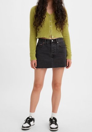 Kvinde iført en grøn, lodden kort cardigan, sort denim mininederdel, hvide sokker og sorte og hvide sneakers, stående mod en ensfarvet baggrund.