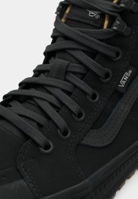 Black high-top sneaker featuring a canvas and suede material, thick black laces, subtle logo detail, and a textured rubber sole.