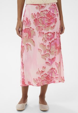 Frau trägt einen rosa Midirock mit Blumenmuster, kombiniert mit beigen Ballerinas, die mit Strasssteinen verziert sind, und steht vor einem schlichten weißen Hintergrund.