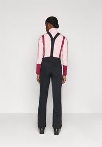 Czarne spodnie narciarskie z regulowanymi szelkami, dopasowany top i zamki błyskawiczne po bokach; w połączeniu z różową i bordową, długostrzałową bluzką o gładkiej teksturze.