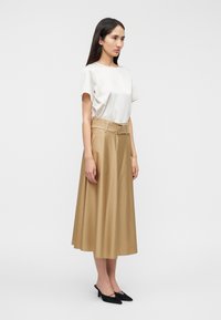 Μπεζ φούστα σε γραμμή Α με ζώνη στη μέση, συνδυασμένη με ανοιχτό κρεμ κοντομάνικο τοπ. Μαύρα παπούτσια με μυτερή μύτη ολοκληρώνουν το σύνολο.