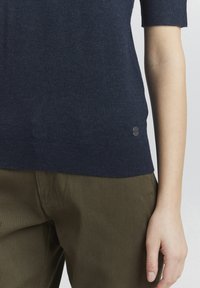 Navy blue knitted short-sleeve sweater with a ribbed hem, paired with olive green pants, featuring a small circular metal logo.