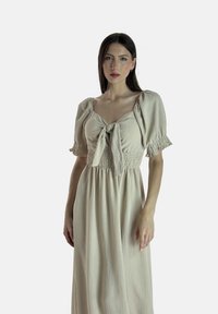 Abito lungo beige con scollo quadrato, maniche a pipistrello corte e un nodo frontale. Il corpetto presenta un design smock, creando texture.