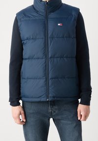 Mörkblå pufferväst med hög krage, framdragkedja och horisontell sömnad. Logotyp patch på vänster bröst. Bärs över en mörk tröja.