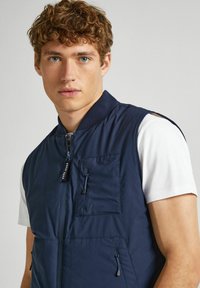 Marinblå väst med dragkedja fram, ribbat krage och flera fickor. Tillverkad av ett slätt tyg med lätt design och en matt finish.