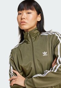 Maslinasto zelena Adidas trenirka s bijelim trakama, cijelim zatvaračem, visokim ovratnikom, glatkom teksturom i malim logotipom na prsima.