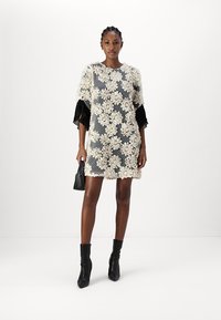 Lace blomsterkjole har en grå bakgrunn med elfenbensfargede blomster, svarte frynsete ermer og rett snitt. Svarte ankelsko kompletterer antrekket.