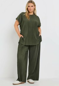 Grön plisserad topp med korta ärmar och sidofickor, matchad med vida byxor i samma material och färg, visad med vita skor.