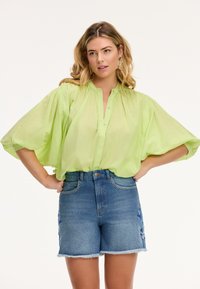 Blusa trasparente verde lime con collo alto e maniche ampie, abbinata a pantaloni shorts in denim a vita alta con orli sfilacciati e ricami laterali.
