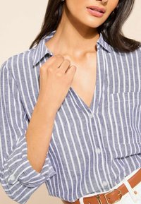Kék és fehér csíkos gombos ing, zsebbel, felhajtott ujjakkal és V-nyakú kialakítással. Simogató anyag, laza szabás.