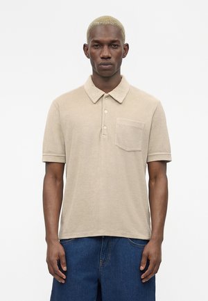 Homme aux cheveux blonds courts portant un polo beige à manches courtes avec une poche sur le devant et un pantalon en jean bleu sur fond blanc.
