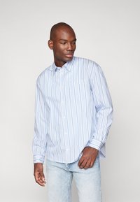 Camisa de manga larga a rayas en azul claro y blanco, con botones en la parte delantera, un bolsillo en el pecho, corte relajado y un sutil detalle de textura en la tela.