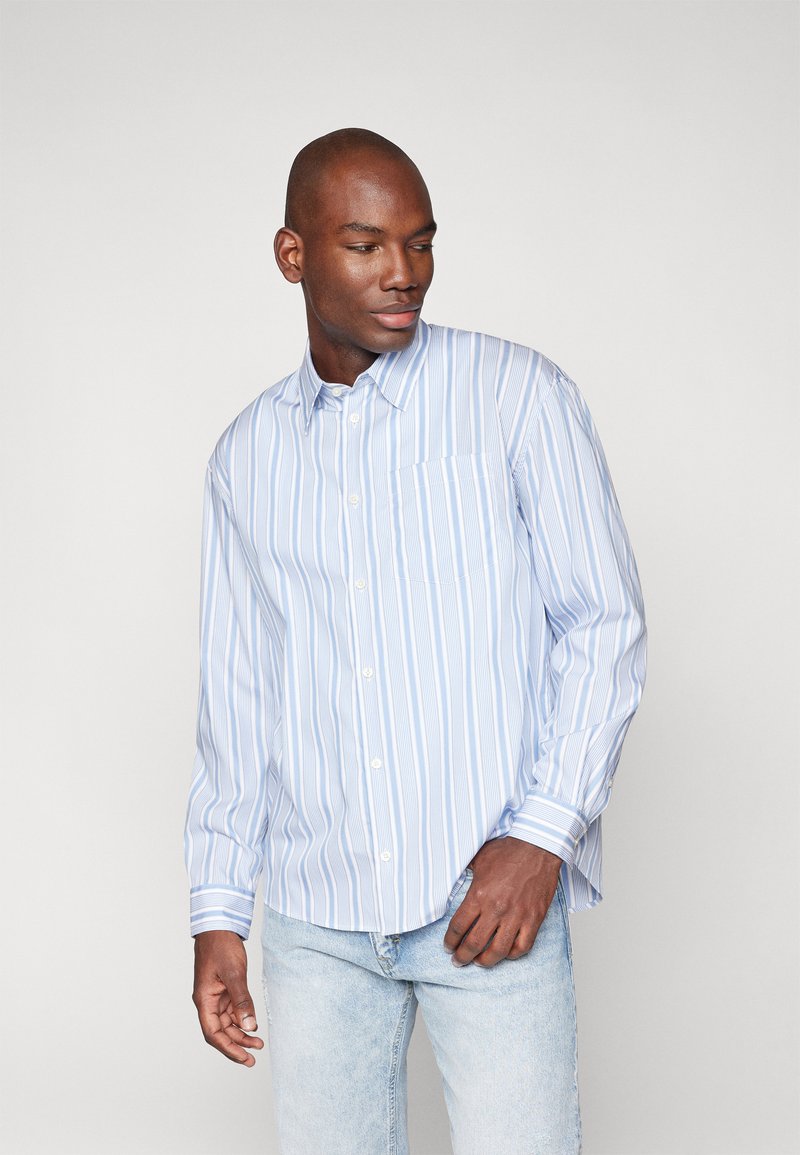 Camisa de manga larga a rayas en azul claro y blanco, con botones en la parte delantera, un bolsillo en el pecho, corte relajado y un sutil detalle de textura en la tela.