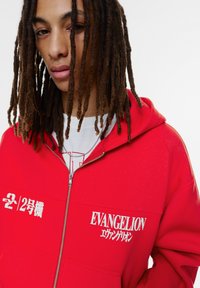 Roter Zip-Hoodie aus weichem Material mit dem Schriftzug "EVANGELION" und grafischem Text in Weiß. Enthält eine geräumige Kapuze und eine lockere Passform.