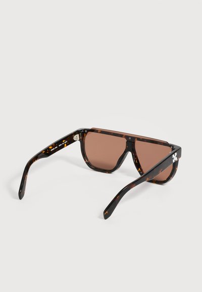 Lunettes de soleil avec de grandes lentilles teintées marron et des montures motif écaille noire et marron posées sur une surface blanche.