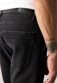 Pantaloni din denim negru cu o croială slim, având două buzunare la spate și o etichetă din piele. Materialul texturat prezintă o ușoară decolorare în anumite zone.