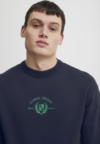 Mørkeblå bomullsgenser med ribbestrikket rund hals. Har grønn brodert tekst "CASUAL FRIDAY" og et logo med laurbærakenter.