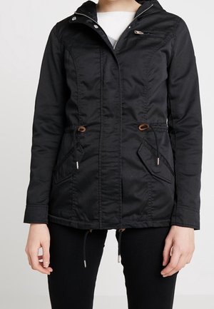 Femme portant une veste noire zippée avec capuche, poches avant à rabat et cordons réglables à la taille et à l’ourlet.
