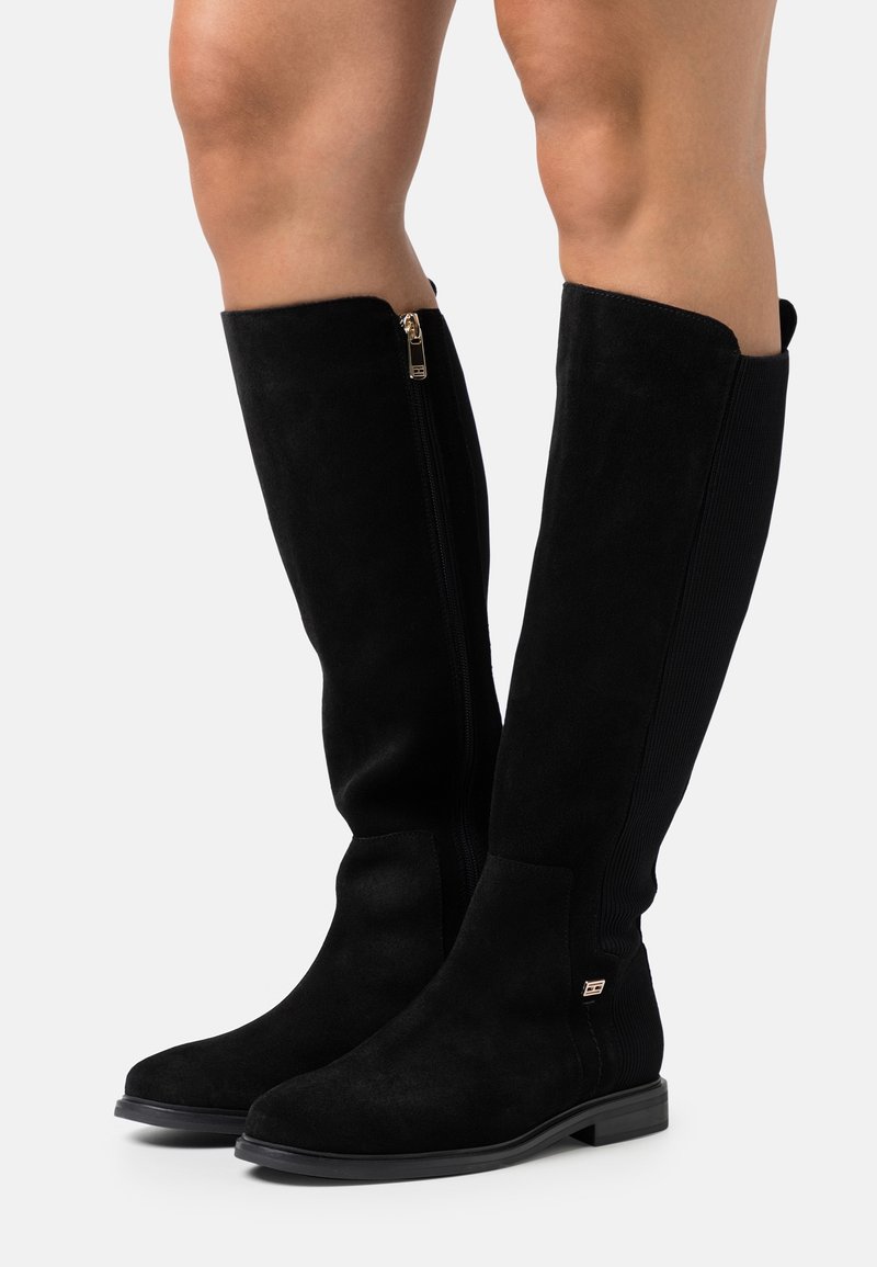 Knee-high black suede boots with a zipper detail and ribbed elastic panels, featuring a flat black sole and subtle metallic accents.