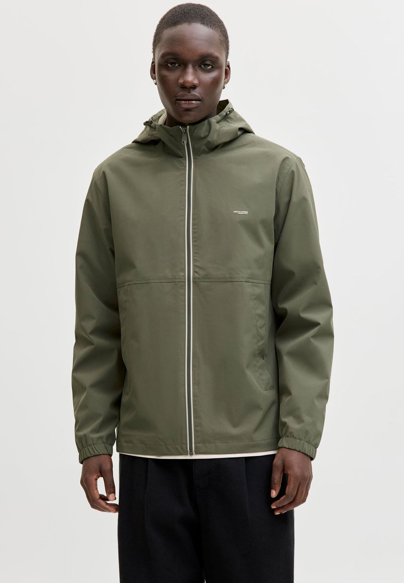 Młoda osoba z krótkimi włosami, ubrana w oliwkowo-zieloną kurtkę z kapturem i czarne spodnie, stojąca na jasnym, jednolitym tle.