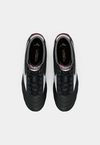 Crne nogometne kopačke s teksturiranim crnim gornjim dijelom, bijelim bočnim prugama i crnim vezicama. Imaju podstavljeni ovratnik i crveni naglasak na jeziku.