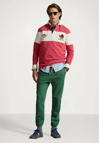 Mężczyzna noszący okulary przeciwsłoneczne, czerwono-białą koszulę rugby z logotypami, jasnoniebieską koszulę pod spód, zielone spodnie dresowe i niebieskie trampki, stoi z jedną ręką w kieszeni.