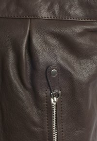 Bruine leren tas met een zachte textuur, voorzien van een rits en drukknopdetails. Gestikte accenten verbeteren het algehele ontwerp.