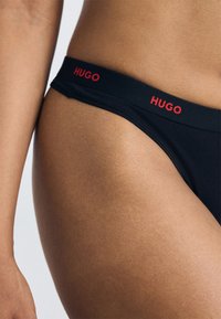 Juodos maudymosi kelnaitės su lygiu paviršiumi ir raudonu "HUGO" logotipu ant juosmens, pasižyminčios minimalistiniu dizainu ir žemu kirpimu.