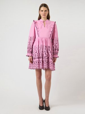 Donna in piedi che indossa un vestito rosa con maniche lunghe, volant, ricamo floreale nero e scarpe nere con tacchi alti su sfondo bianco.