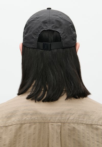 Osoba s crnom kosom do ramena, koja nosi sivi podesivi bejzbol kapu i bež majicu, okrenuta leđima na bijeloj pozadini.