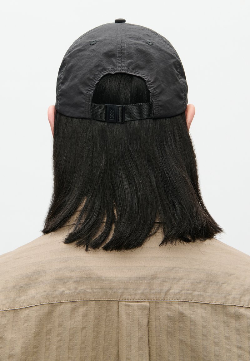 Persona con capelli neri di lunghezza medio-lunga che indossa un cappellino da baseball grigio regolabile e una camicia beige, di spalle su uno sfondo bianco.