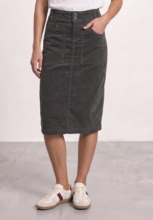 Femme portant une jupe sombre mi-longue avec des poches avant et des baskets blanches décontractées avec des rayures bordeaux.