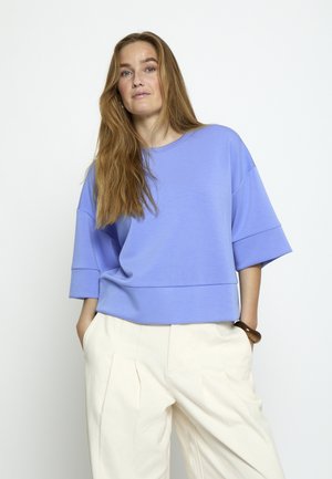 Frau mit langem Haar, die ein lockeres hellblaues Oberteil und hoch taillierte weiße Hosen trägt, mit den Händen in den Taschen vor einem einfarbigen Hintergrund.