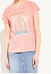 Lyserød t-shirt med korte ærmer og et struktureret mønster, der har et kunstnerisk dørprint. Bæres med en lys blå denimnederdel med knapper.