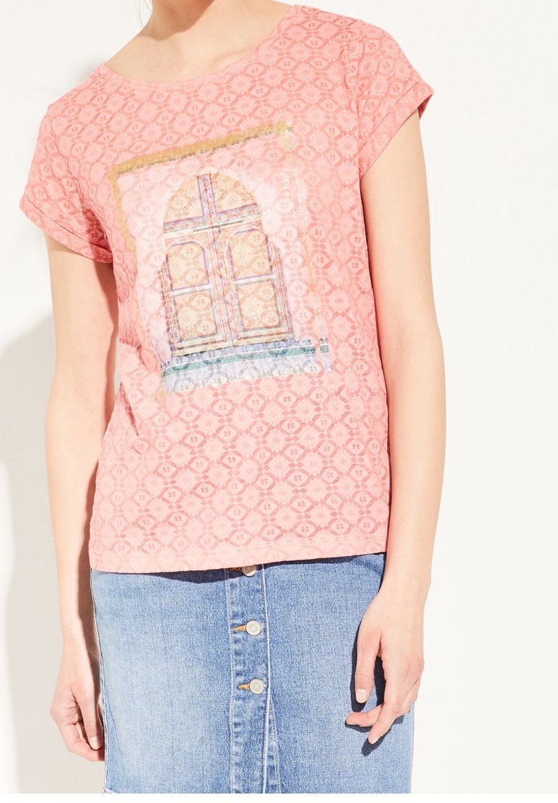 Lyserød t-shirt med korte ærmer og et struktureret mønster, der har et kunstnerisk dørprint. Bæres med en lys blå denimnederdel med knapper.