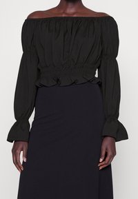 Top noir à épaules dénudées avec des manches longues froncées, un ourlet volanté et une taille élastique. Le tissu a une texture lisse et mate.
