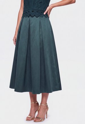 Woman wearing a dark green pleated midi skirt, a dark lace top, and metallic ankle-strap high-heeled sandals, standing with one hand on her hip.