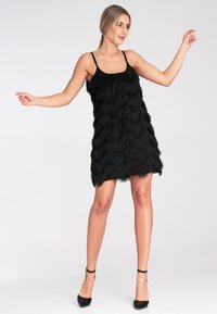 Schwarzes Fransen-Kleid mit geschichteten, wellenförmigen Texturen, Spaghettiträgern und einem durchsichtigen Saum. Kombiniert mit schwarzen High Heels.
