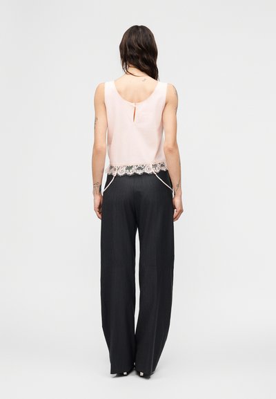 Γυναίκα με μακριά σκούρα μαλλιά που φοράει αμάνικο ανοιχτό ροζ τοπ με δαντέλα στο τελείωμα και φαρδιά σκούρα παντελόνια, στέκεται γυρισμένη από πίσω.