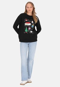 Sudadera negra con ilustraciones coloridas de temática navideña, cuello redondo, puños y dobladillo acanalados. Combinada con vaqueros azul claro y zapatillas blancas.