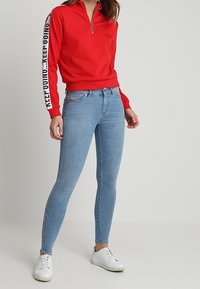 Sudadera roja con cremallera y texto blanco en las mangas, combinada con jeans ajustados de color azul claro y zapatillas blancas. Tela suave, diseño entallado.