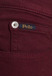 Le tissu en denim bordeaux présente un bouton en métal et une étiquette bleu foncé avec "Polo" en fil doré sur la poche arrière.