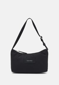 Musta nailoninen olkalaukku vetoketjulla, suorakaiteen muotoinen, säädettävä hihna ja Calvin Klein -logopatchi edessä.